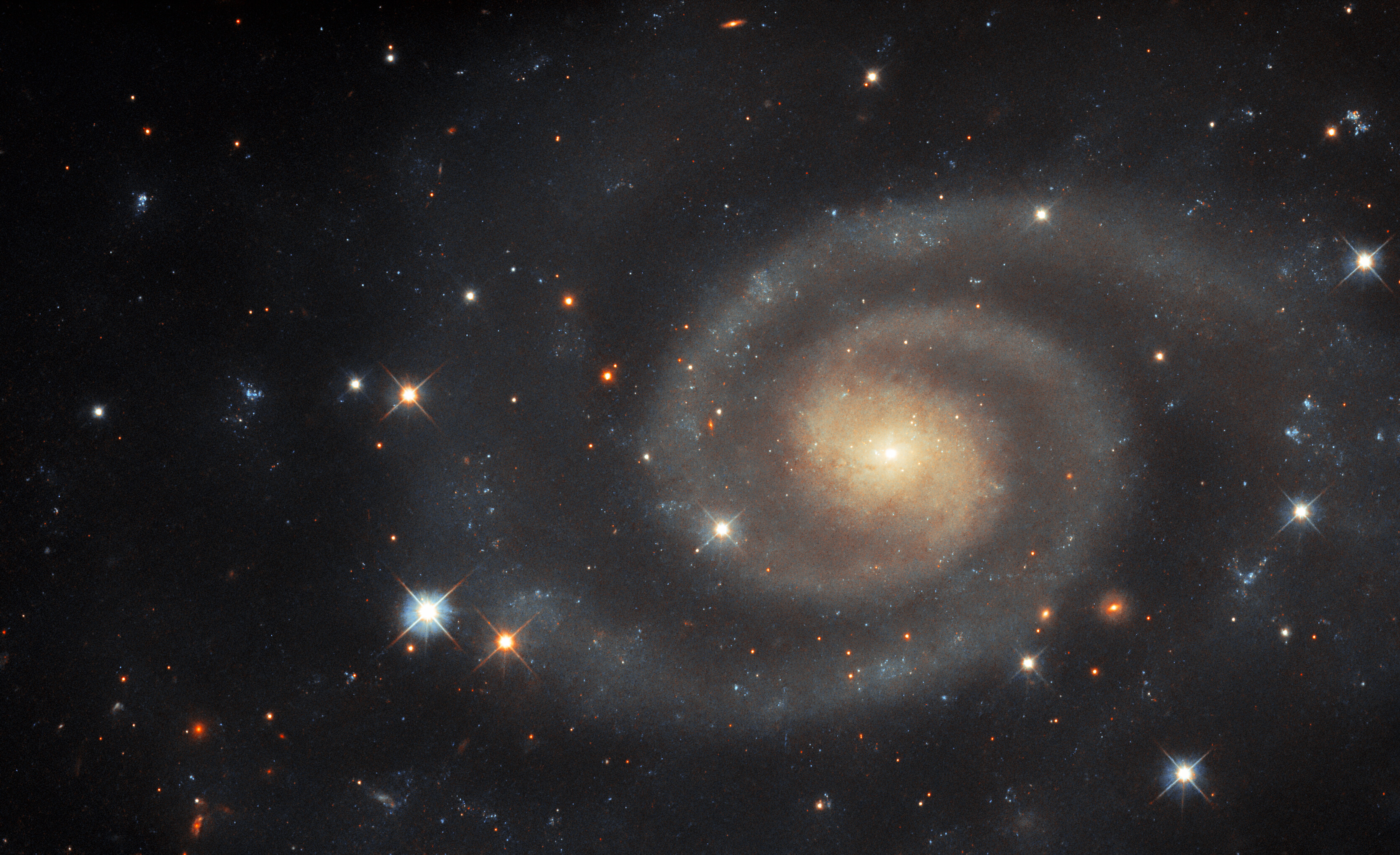 A spiral galaxy, with two prominent, tightly wound arms around the brighter core. The arms disperse into a wide halo of stars and dust at their ends, giving the galaxy an oval shape. A number of bright, foreground stars appear to flank the galaxy, each holding the signature cross of diffracting light. The image also holds a few distant background galaxies.