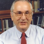 John Bahcall looks at the camera in front of a bookshelf. He wears a light gray shirt, red tie, and glasses.