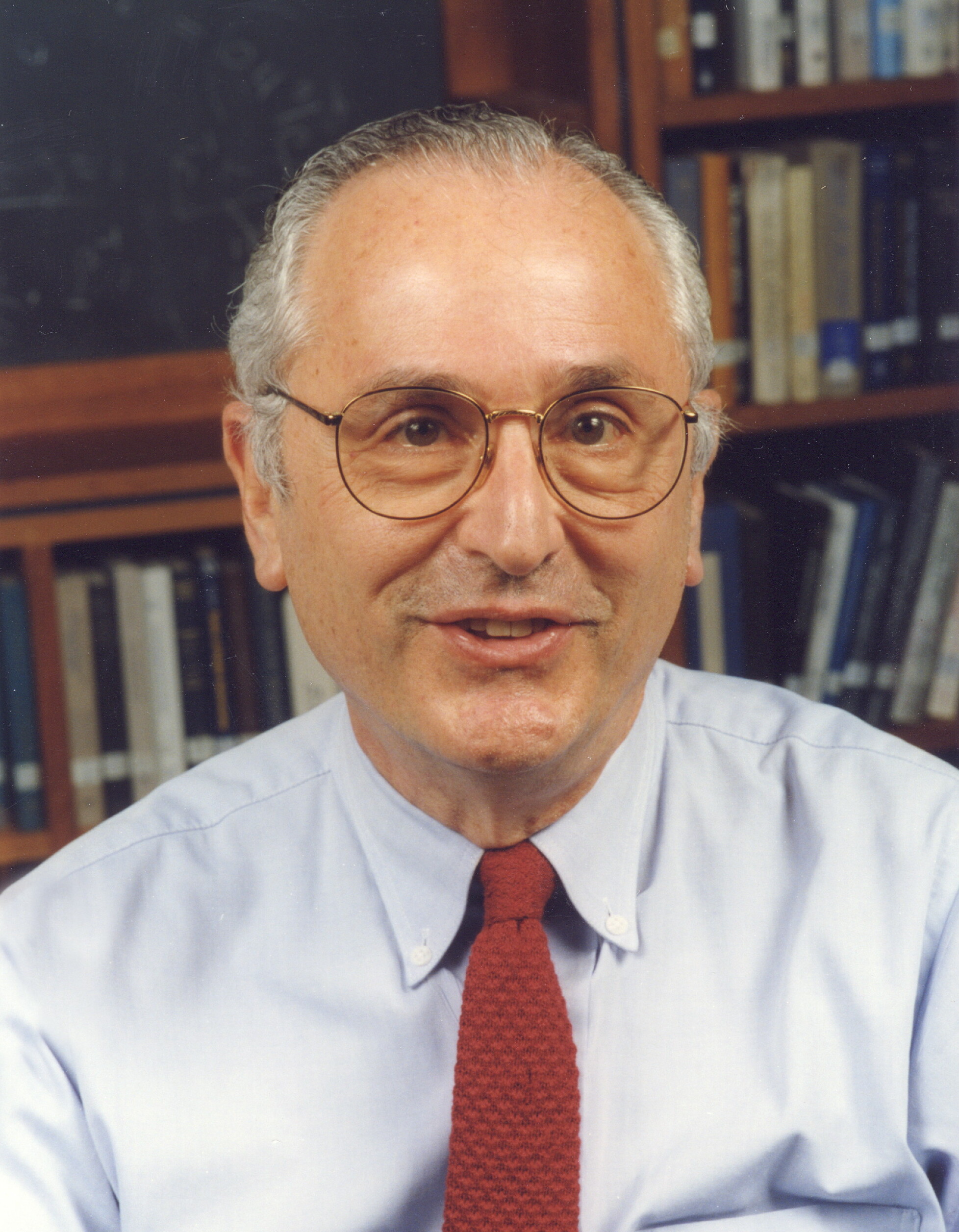 John Bahcall looks at the camera in front of a bookshelf. He wears a light gray shirt, red tie, and glasses.