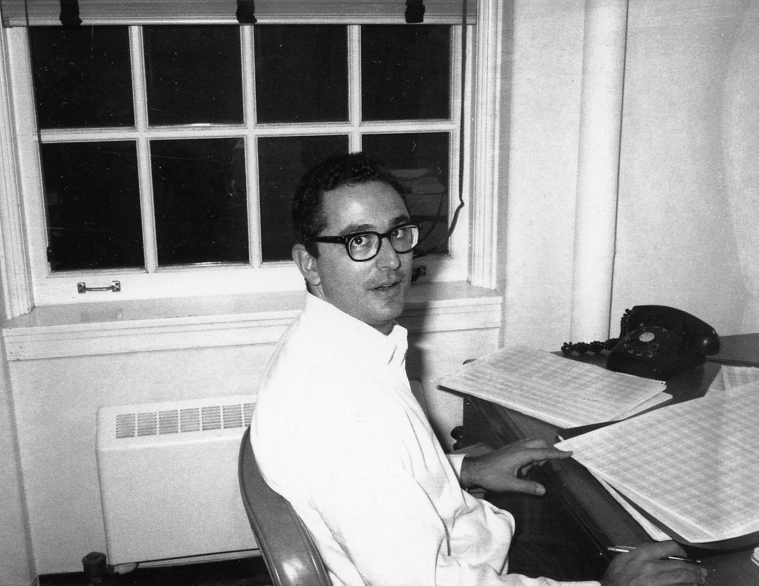 Astronomer John Bahcall sits at a desk facing toward the right. His head is turned toward the camera.