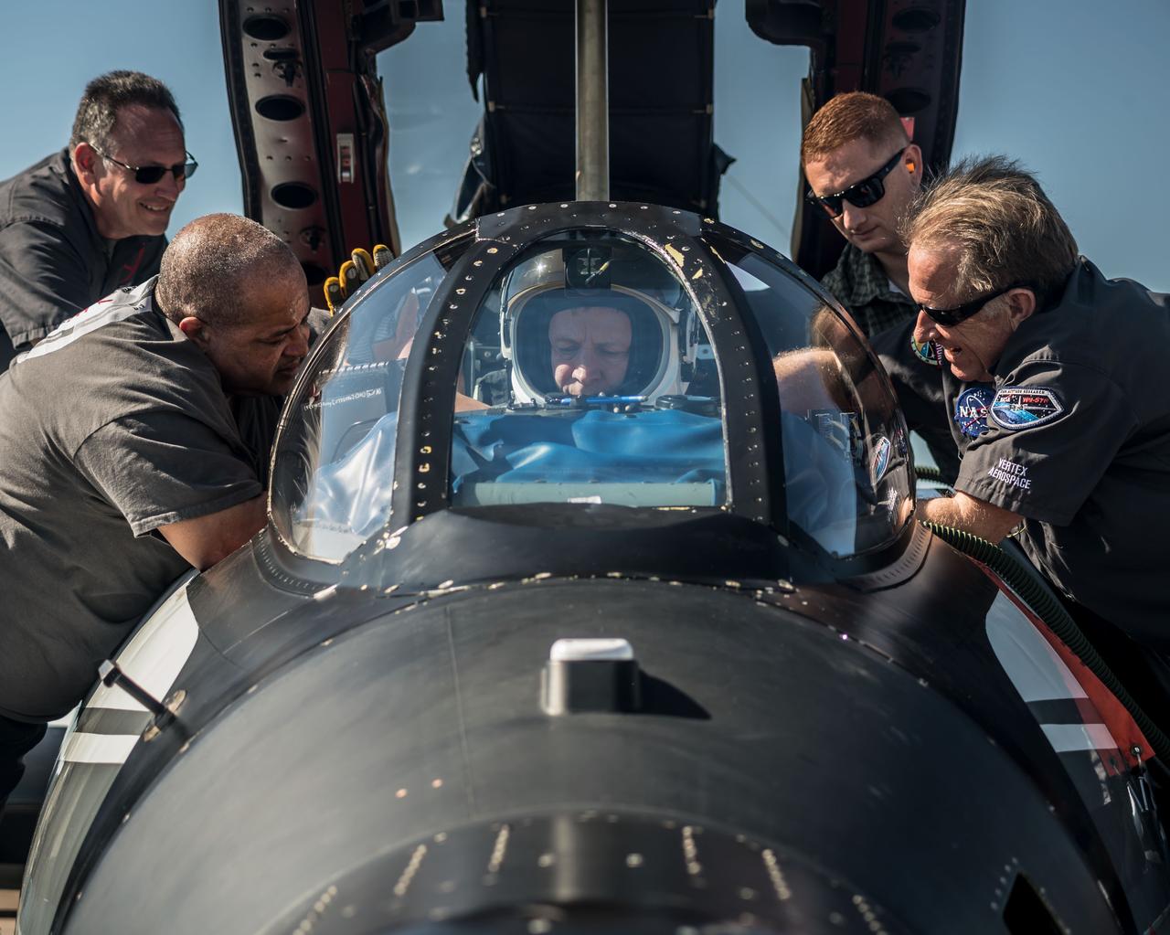 A pilot sits in the cockpit of a small aircraft. The are four people – two on each side of him – working to get the pilot situated.