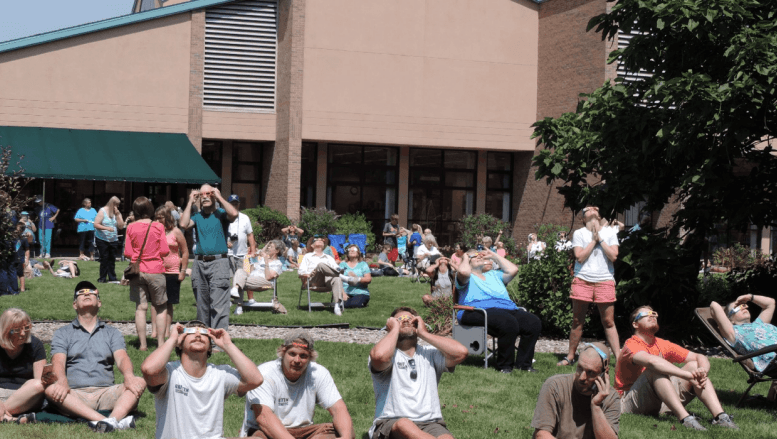 People gather in a field wearing eclipse glasses and looking up at the sky to observe a solar eclipse.