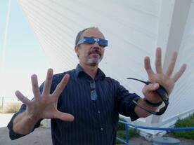 Photo of a man outside wearing protective eyewear for the eclipse and holding his hands to the camera with his fingers spread out.