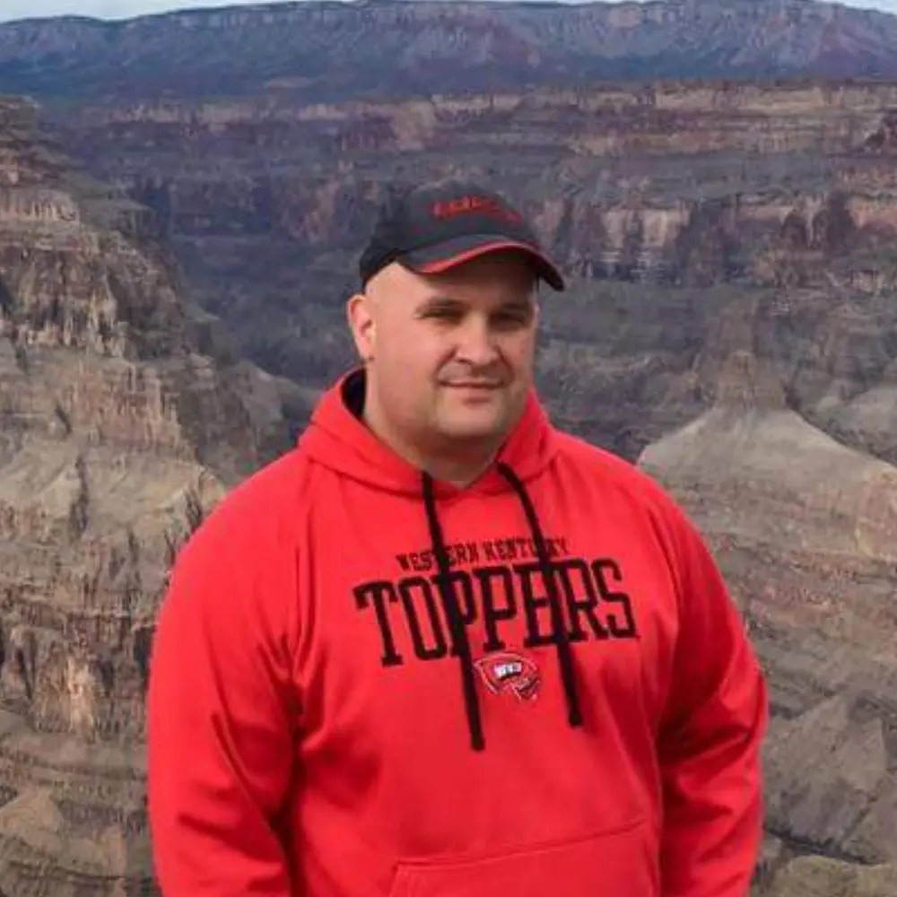 Portrait photo of a man wearing a black ball cap and a bright red sweatshirt standing with the Grand Canyon in the background.