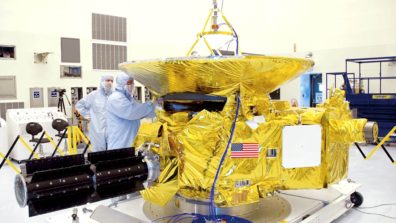 Two men work on a grand piano-sized spacecraft wrapped in a gold insulating blanket. A large cylinder, a radioisotope thermoelectric generator, sticks out of the side.