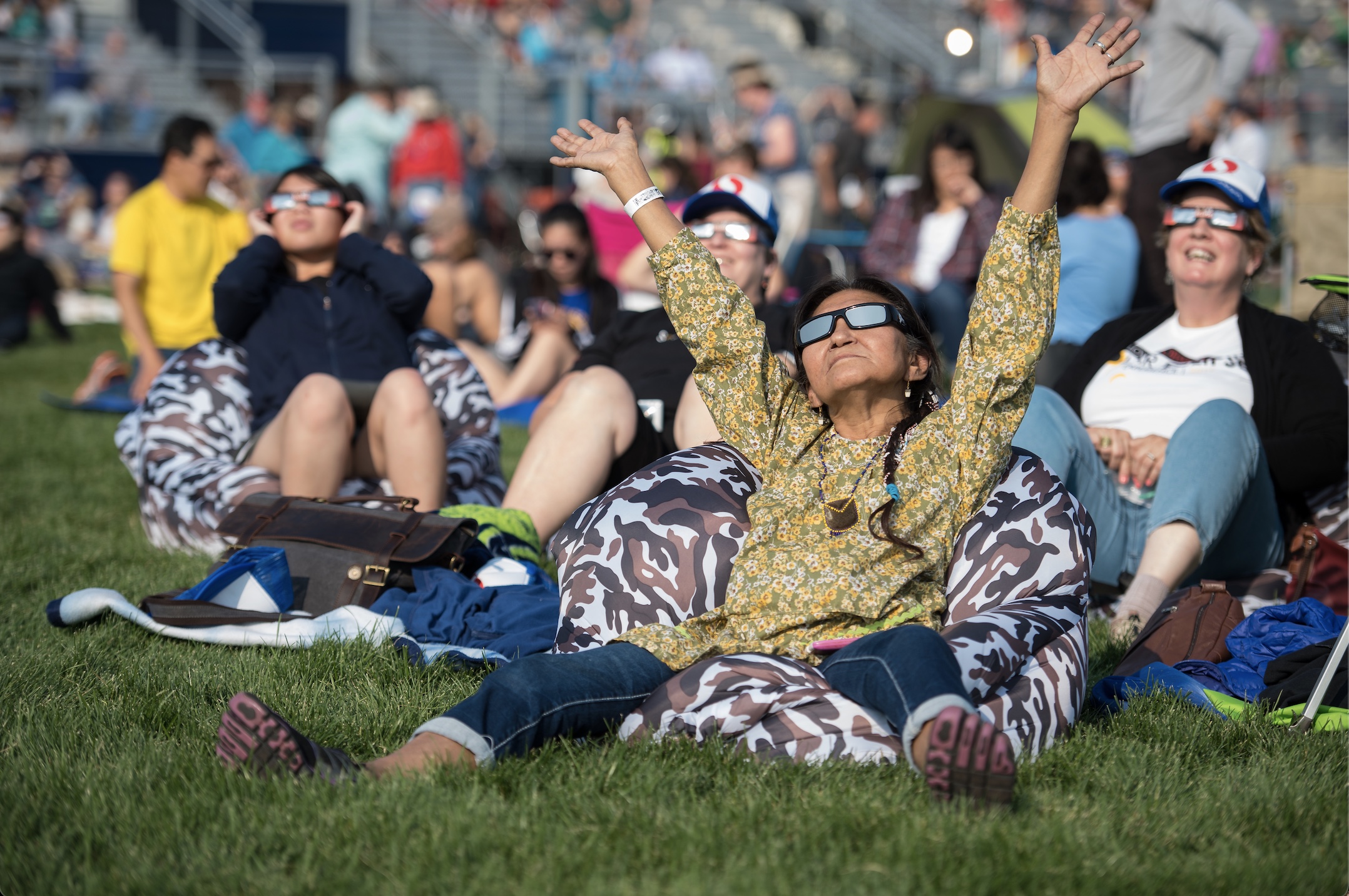 A woman wearing eclipse glasses gazes up and stretches her hands and arms to the sky in celebration of such an amazing experience. Other people can be seen in the background, also wearing eclipse glasses and enjoying the view.