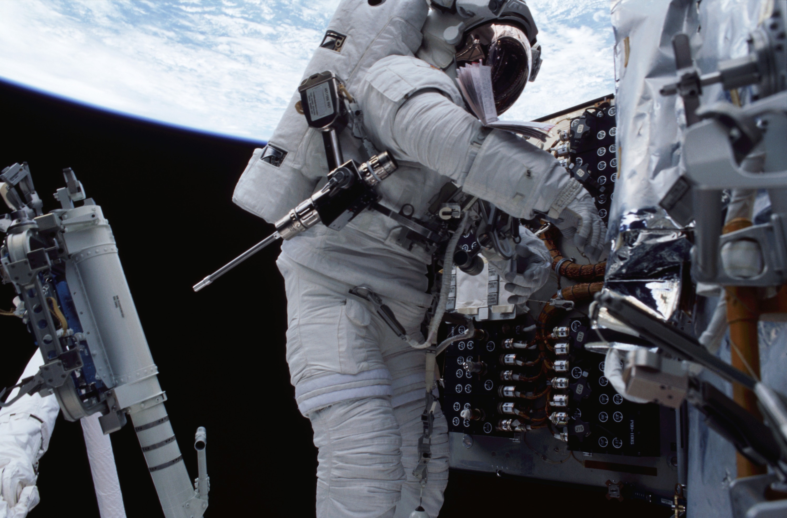 An astronaut in a spacesuit has a long tool that resembles a drill attached to his belt as he works on the Hubble space Telescope. Earth is visible in the background.