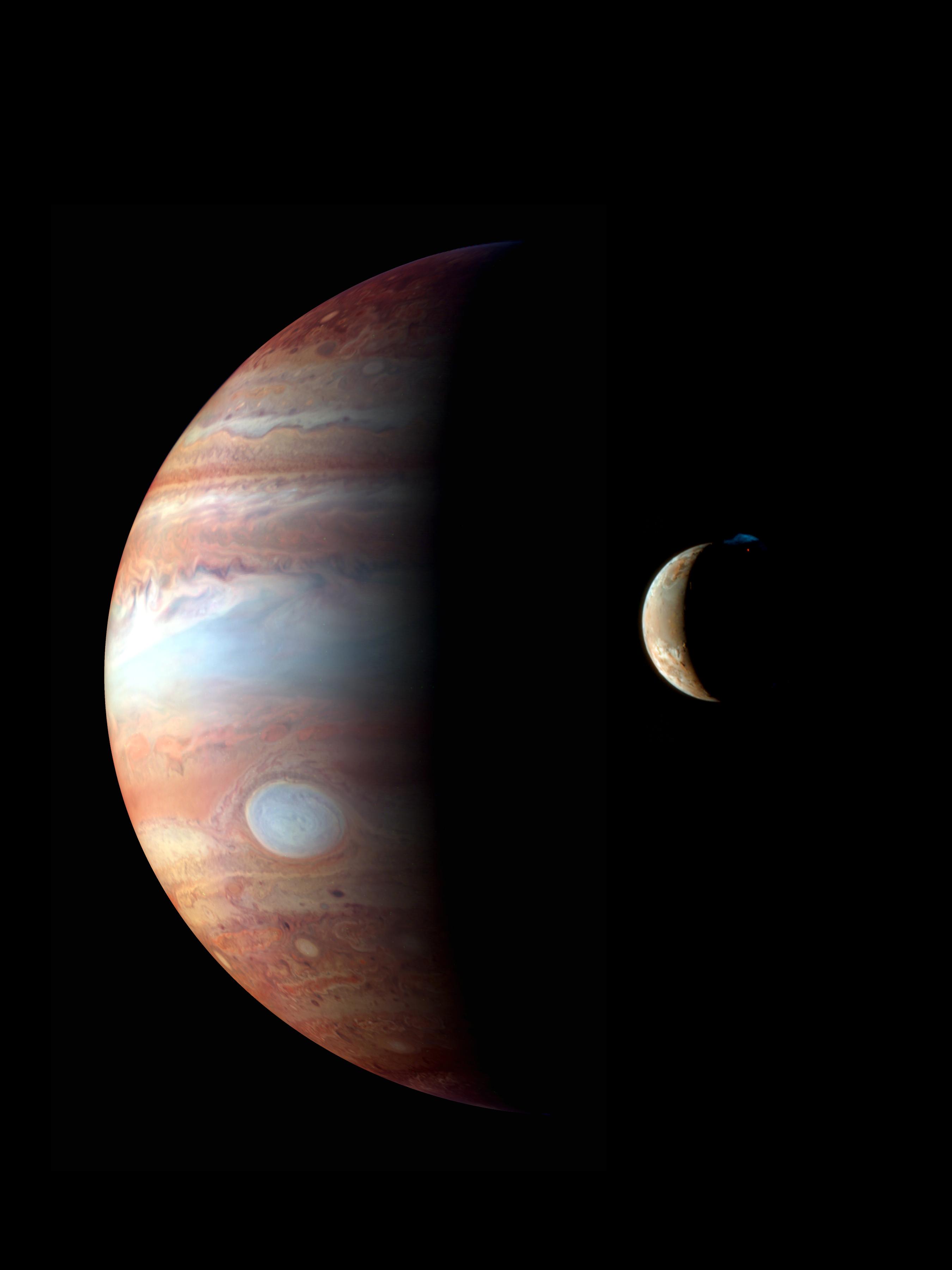 Giant Jupiter is half illuminated by the Sun and its moon Io is a crescent in the foreground. A small glowing volcanic plume is erupting from Io.