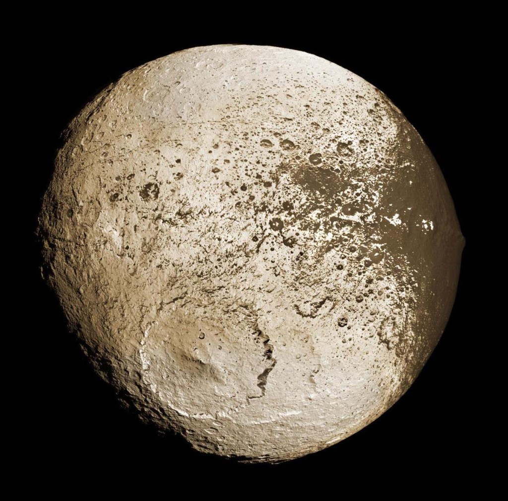 The full disc view of this moon is dominated by a massive impact crater and a dark splatter of terrain hat stands out from the moon's surface.