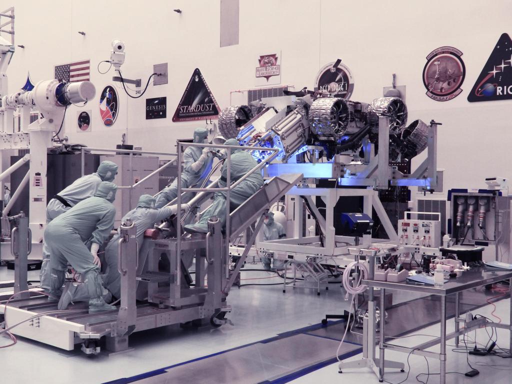 Five engineers look intently at the upside-down Perseverance Rover. They are examining the rover's Radioisotope Thermoelectric Generator.