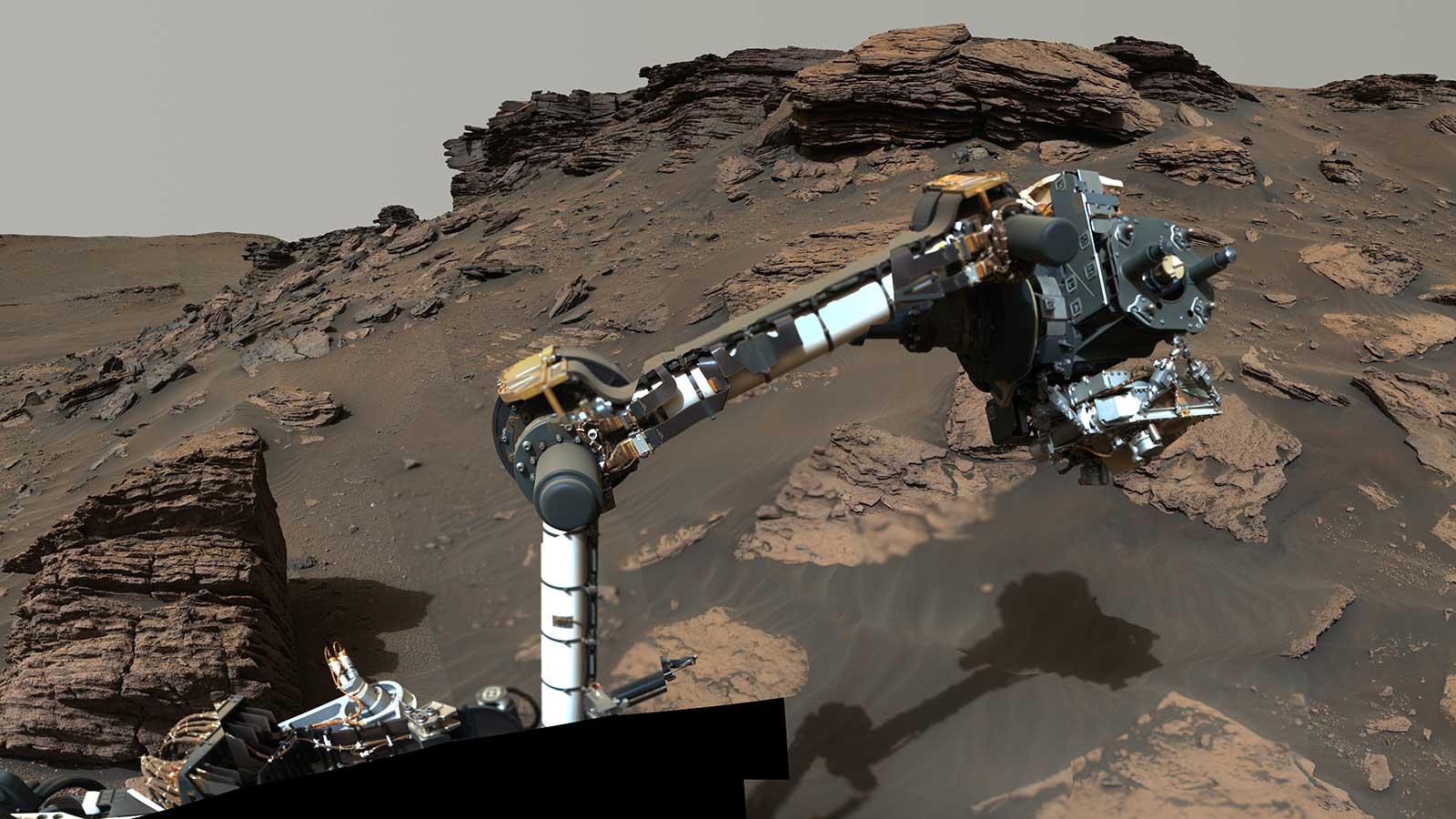 A robotic arm laden with science instruments extends toward a rocky outcrop on Mars.