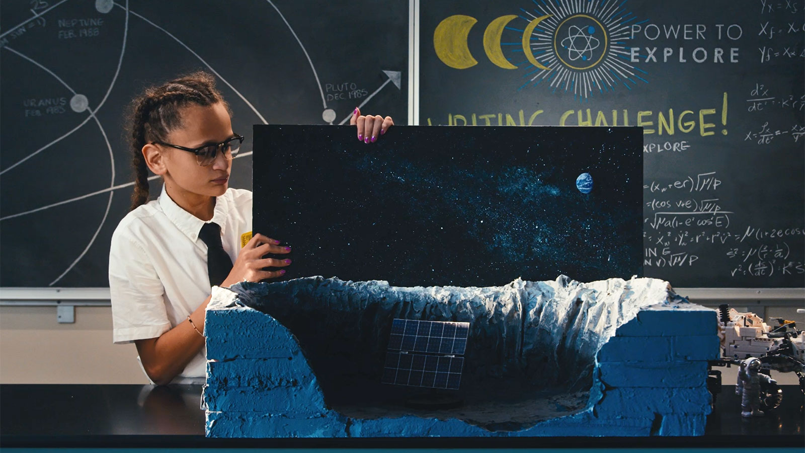 A middle-school-aged girl dressed like an old school NASA engineer (short-sleeved white shirt and black tie) displays a model of a moon crater.