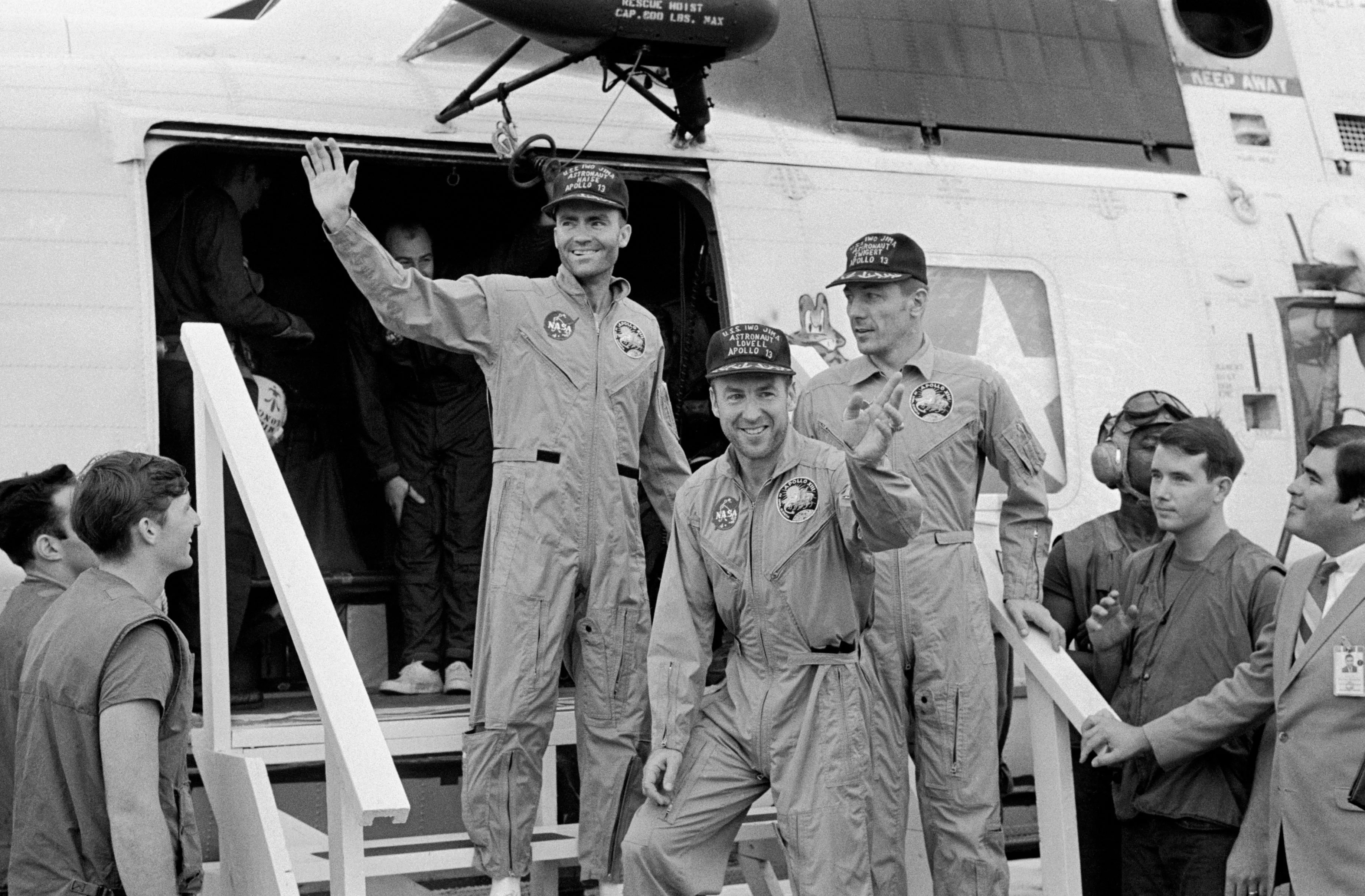 A trio of smiling astronauts step off a helicopter onto the deck of an aircraft carrier in the ocean.