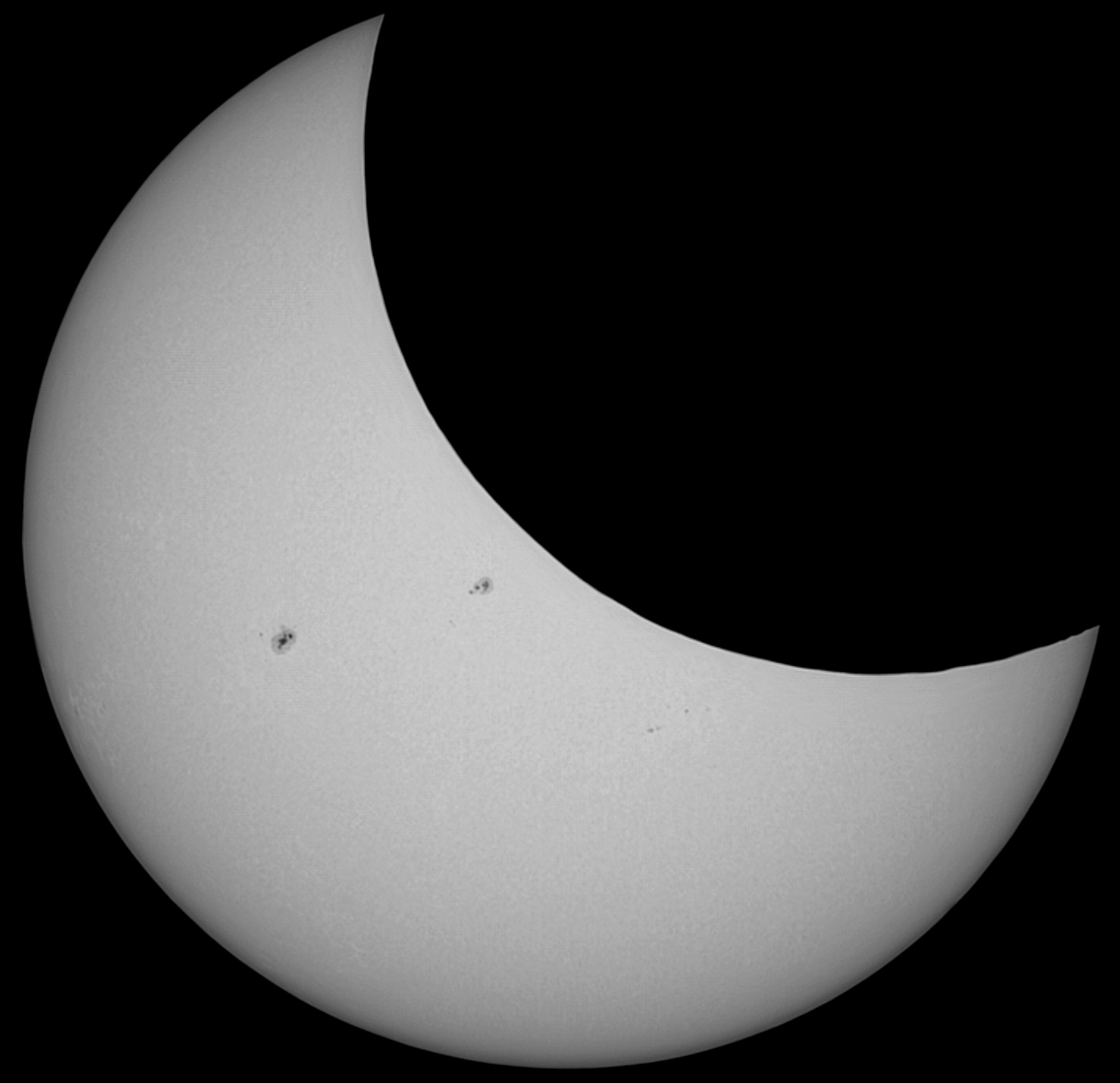 The spherical Sun appears in subtle shades of grey, nearly filling the square frame. The upper right third of the Sun is eclipsed by the Moon, which appears in purest black. There are two dark smudges on the Sun, one in the middle and a slightly larger one halfway between the left edge and the middle. 