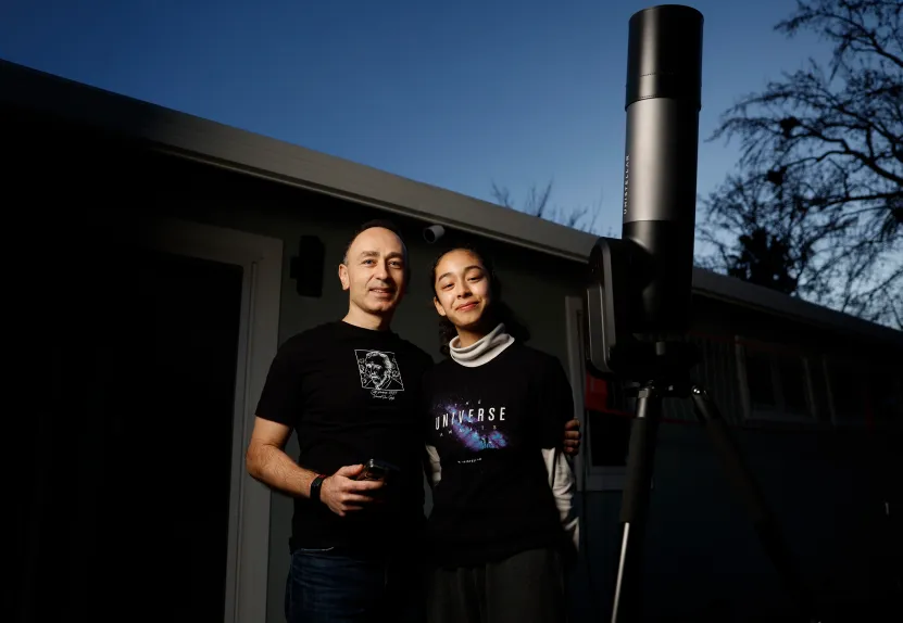 A father and his daughter smile as they pose for a photo next to a large telescope with the sky darkening in the background.