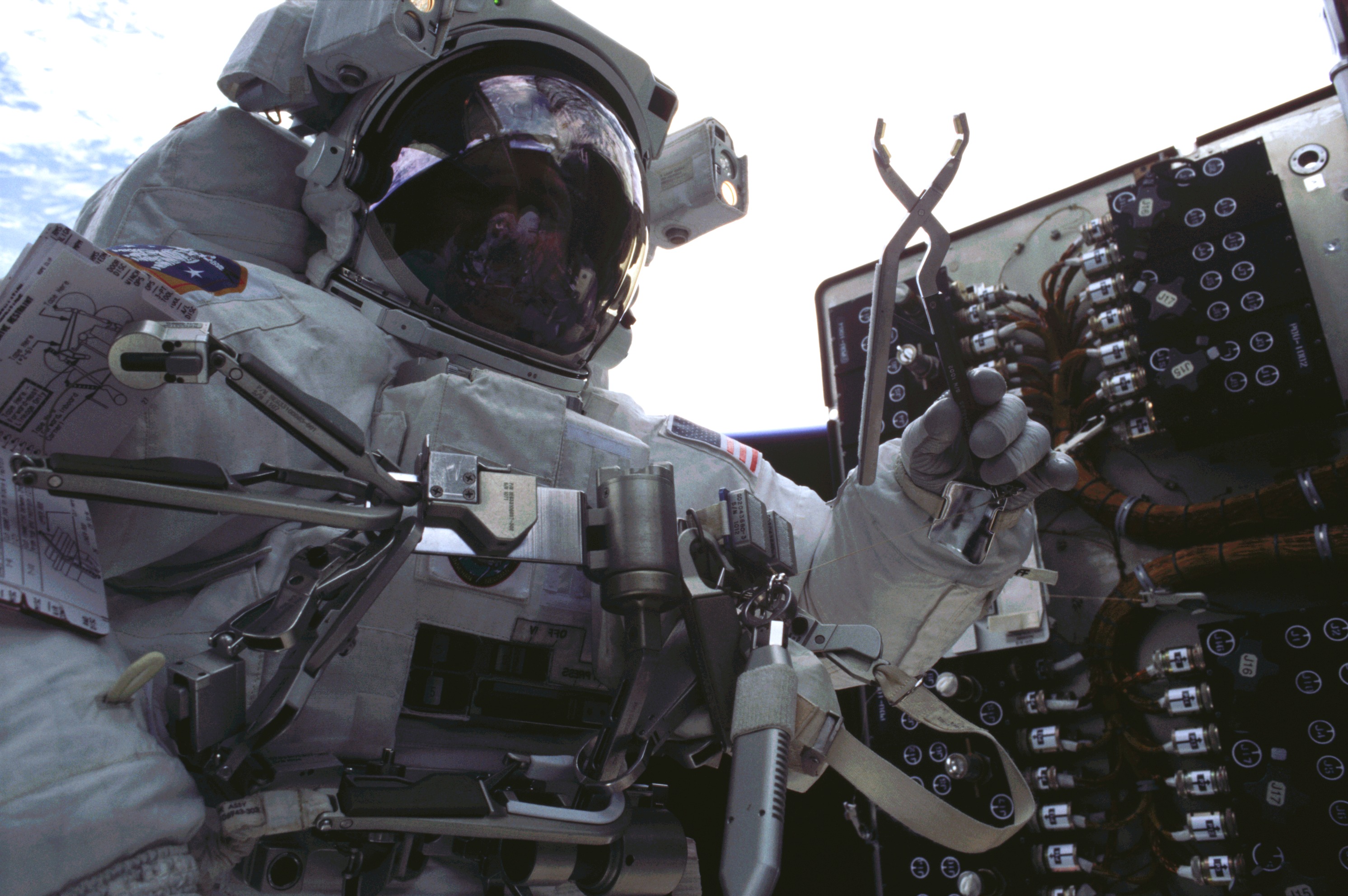 An astronaut in a spacesuit holds aloft a tool resembling a pair of pliers in front of a device covered with wires and connectors. Earth is visible in the background.