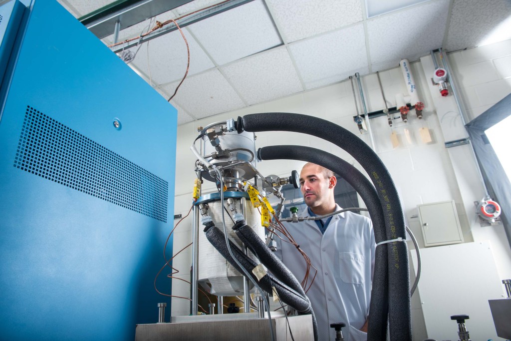 A man in a lab coat is working on a complex machine bristling with tubes, wires and gauges.