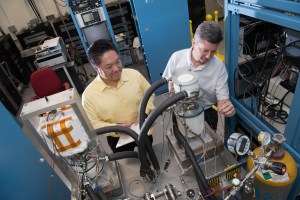 Two men in a room packed with equipment inspect complex system of cylinders, valves and hoses that make up the Stirling Converter.