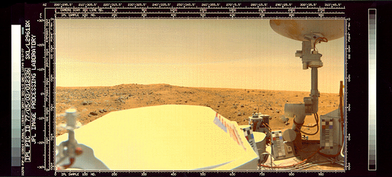 A rocky plain on Mars is visible over the top of the Viking 1 lander. Part of the top deck of the lander, including a large antenna dish, are visible in the foreground.