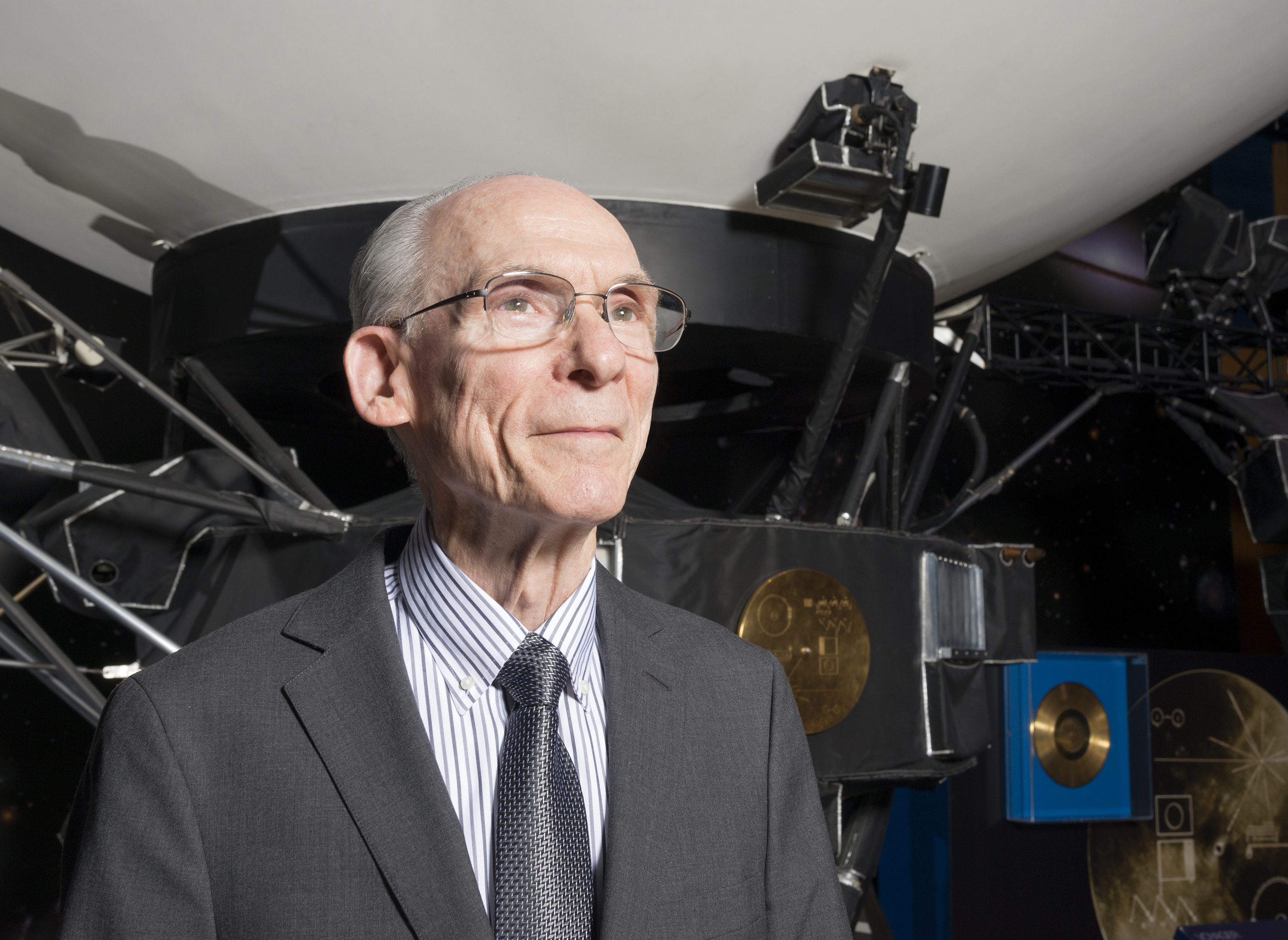 An older man in a coat and tie is standing in front of a full-size Voyager spacecraft model.