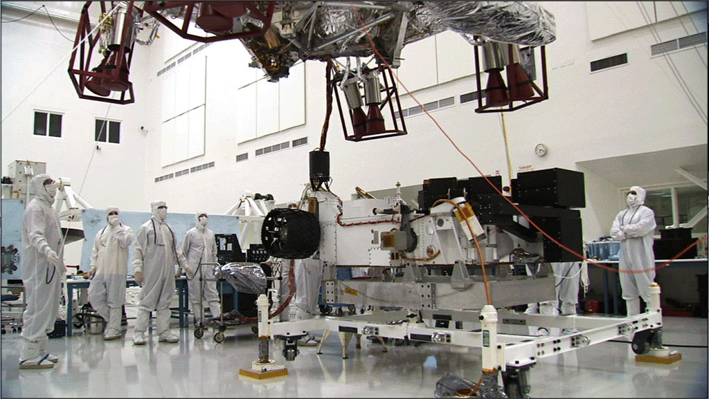 In this image, the car-size rover is in the middle of the picture with several team members surrounding it.  The team members are all dressed in special head-to-toe white suits, called 'bunny suits.'  One team member is holding on to a tether to guide the large insect-like descent stage down on top of the rover.  The descent stage looms high in this image.