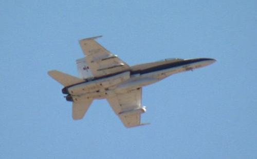 This image shows the underside of a grey F/A-18 jet with dark stripes flying against a blue sky from quite a distance away. Underneath the jet is a pod carrying sensors that measure the jet's temperature, pressure and vibration.