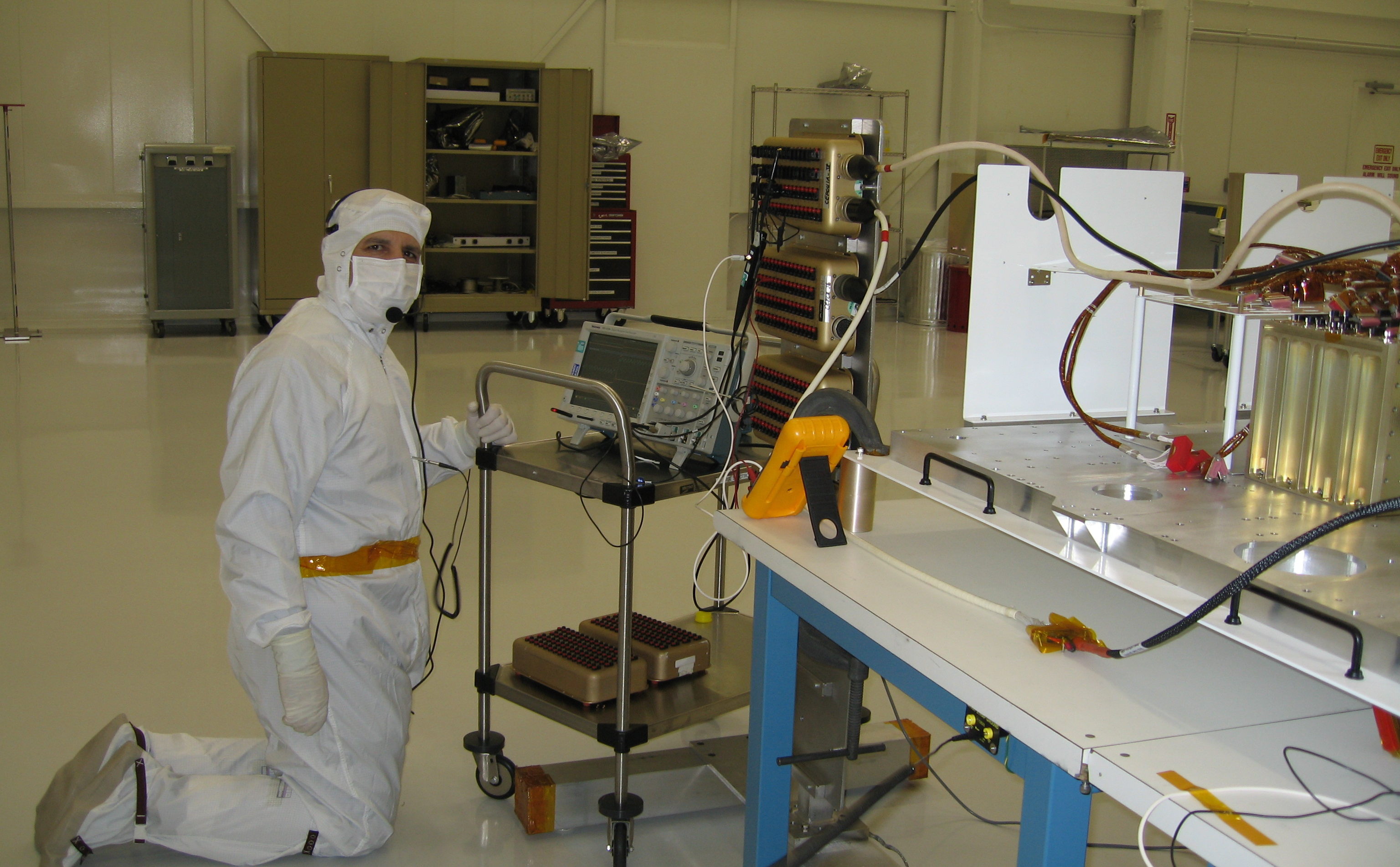 This image was taken inside a large 'clean room,' where ATLO (Assembly, Testing, and Launch Operations) is taking place for the Mars Science Laboratory mission. In the foreground on the right are electronic components and to the left is a man leaning over the components. He is dressed in all white protective clothing, including a face mask and gloves.