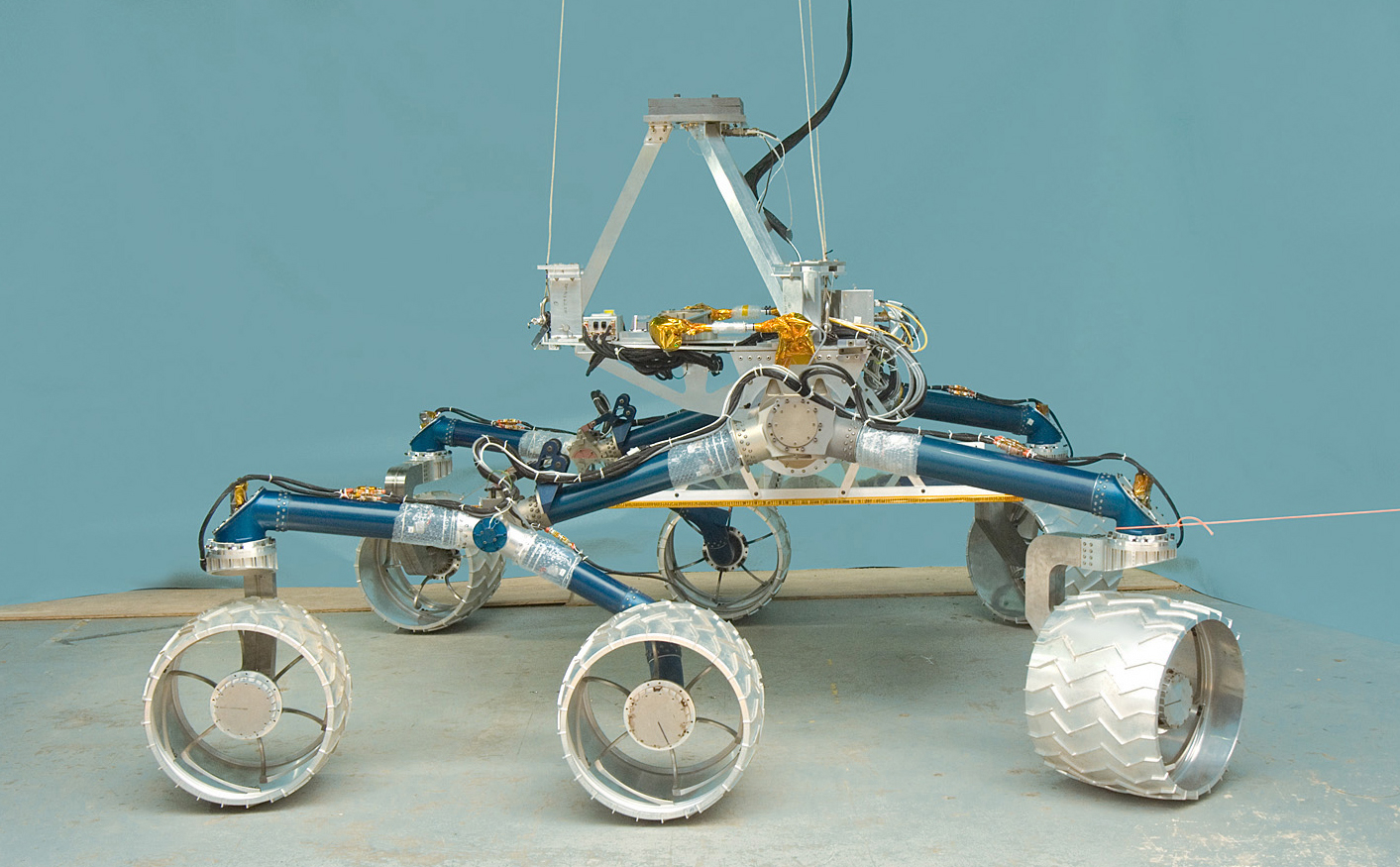 In this image, a large, six-wheeled rover sits on a concrete floor in a laboratory against a light-blue background.  The rover's 'legs,' or mobility system are a medium blue color and the large, cleated wheels are shiny silver.  On top of the mobility system is a silver-colored triangle that represents where the heart and brains of the rover will go when it is complete.