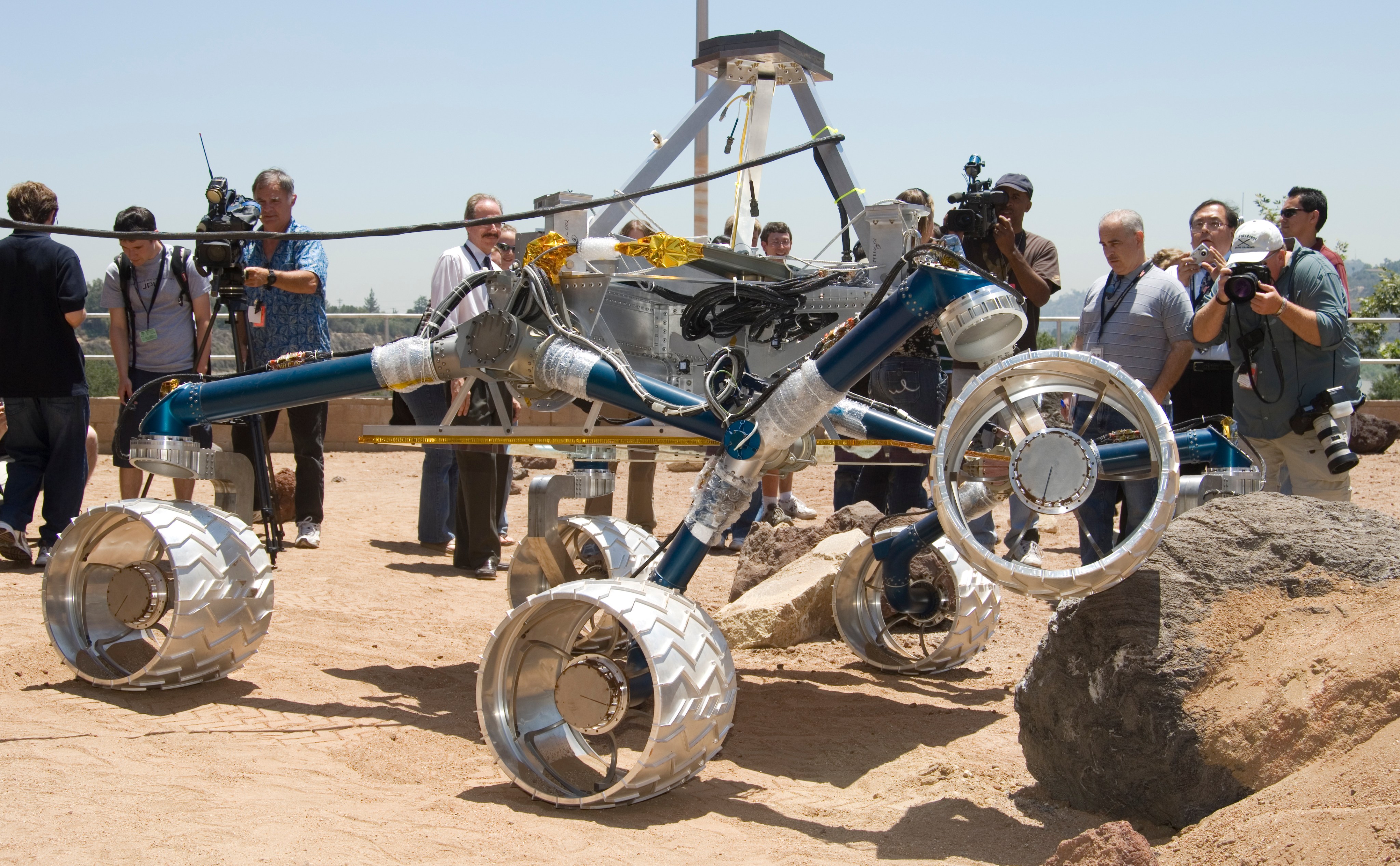 In this image, about a dozen people watch the large Scarecrow rover drive over a grayish orange boulder in an outdoor sandbox.  Scarecrow is about the size of a small compact car.  Its 'legs,' (mobility system) are a vibrant blue color and its large wheels are silver and covered in chevron-like cleats.  Members of the media hold cameras.