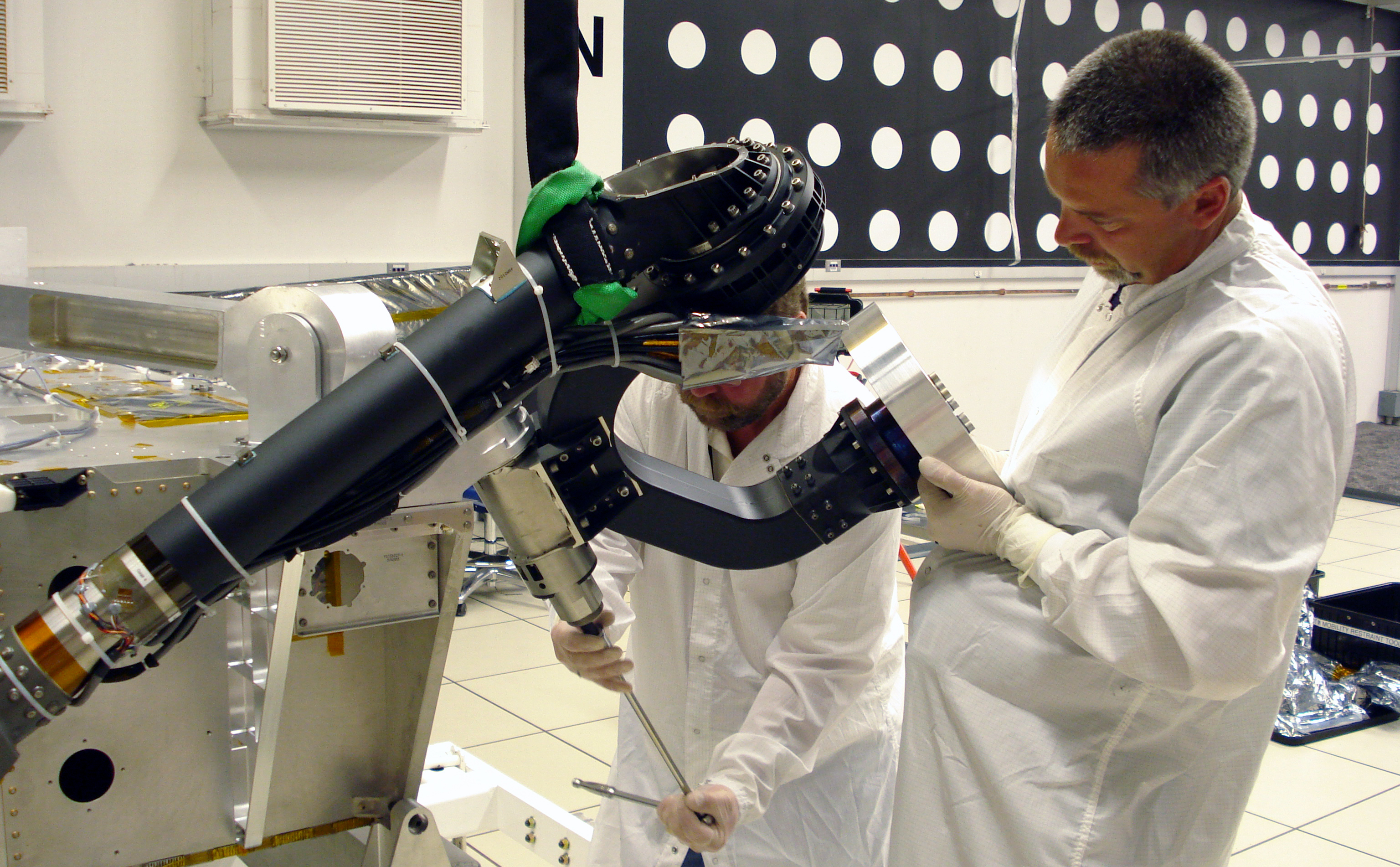 In this image, two technicians from the Mars Science Laboratory team prepare the rover's mobility system for instrumentation tests.  They are both wearing white coats and gloves as they each work on one of the rover's 'legs' in a white-tiled room.
