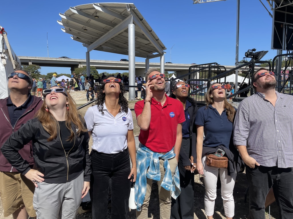 A group of people stand in a line. They're all looking up, wearing eclipse glasses.