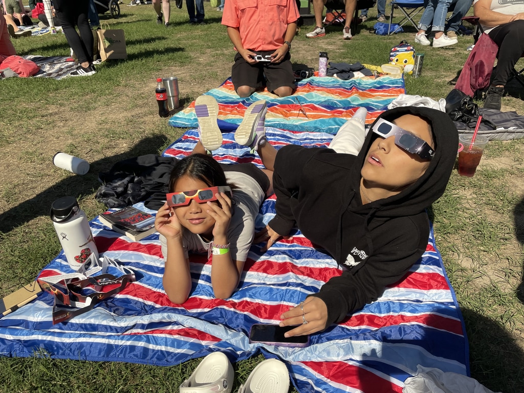 Two children lay on a blanket, looking up, wearing eclipse glasses.