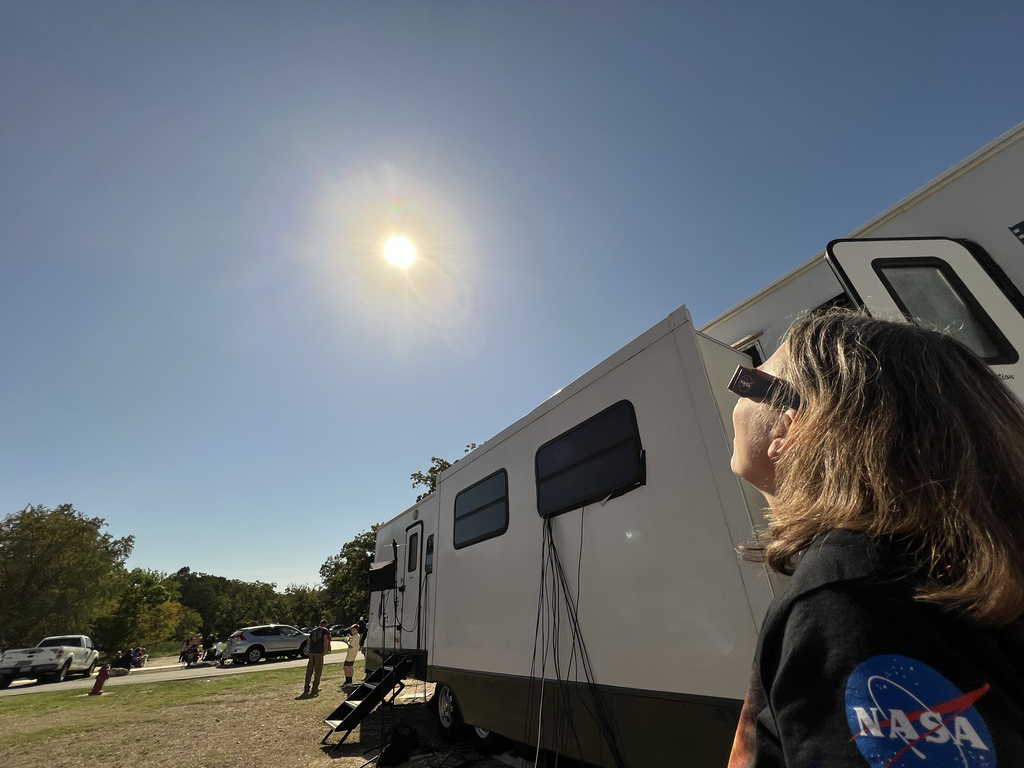 A person standing outside near a trailer, wears eclipse glasses and looks up at an uneclipsed Sun.