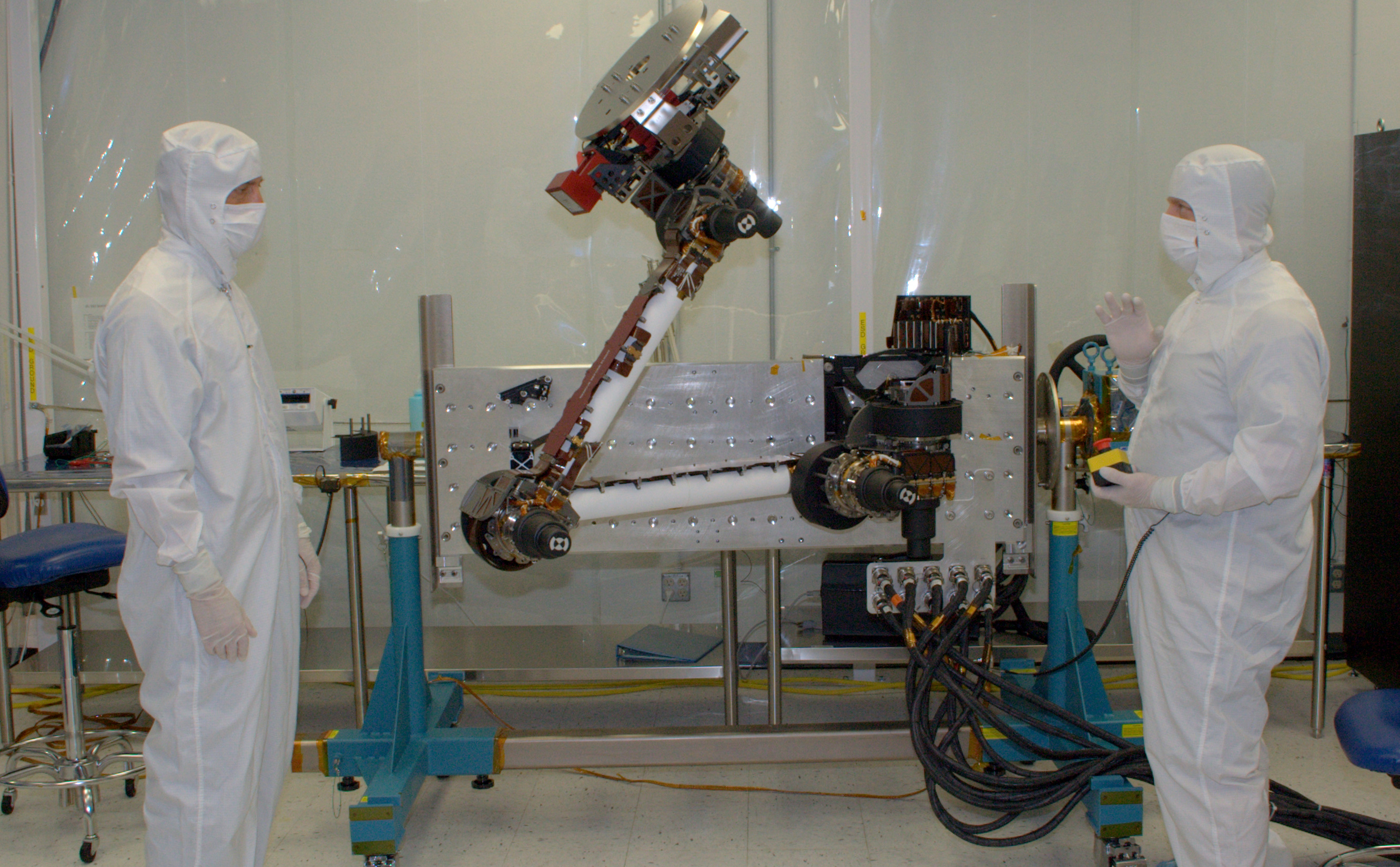 In this photo, two engineers from NASA Jet Propulsion Laboratory and Alliance Spacesystems are standing in a white room on either side of the Mars Science Laboratory rover's robotic arm.  They are dressed in white 'bunny suits,' and are covered from head to toe wearing white face masks to protect the equipment.  The arm is bent at the 'elbow' and raised high in the air about two feet higher than the engineer on the left.  The engineer on the right holds a control box that maneuvers the arm for testing.