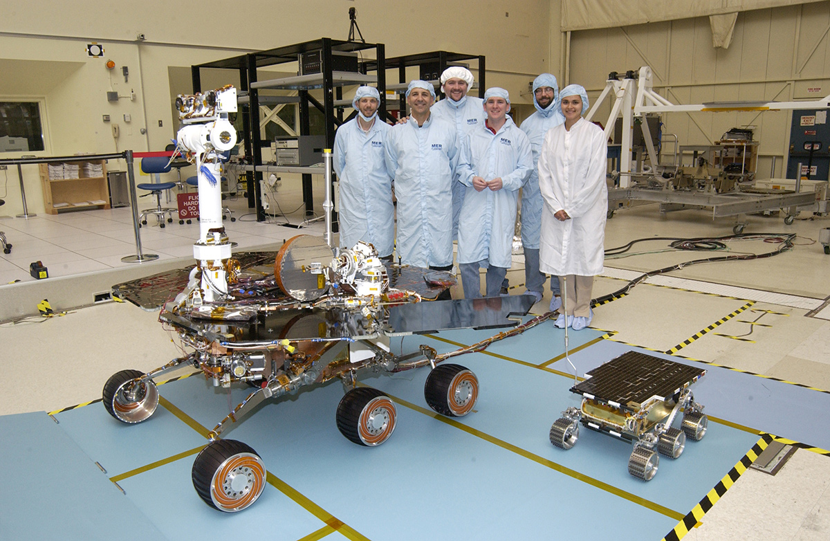 Members of the Mars Exploration Rovers Assembly, Test and Launch Operations team gather around Rover 2 and its predecessor, a flight spare of the Pathfinder mission's Sojourner rover, named Marie Curie.