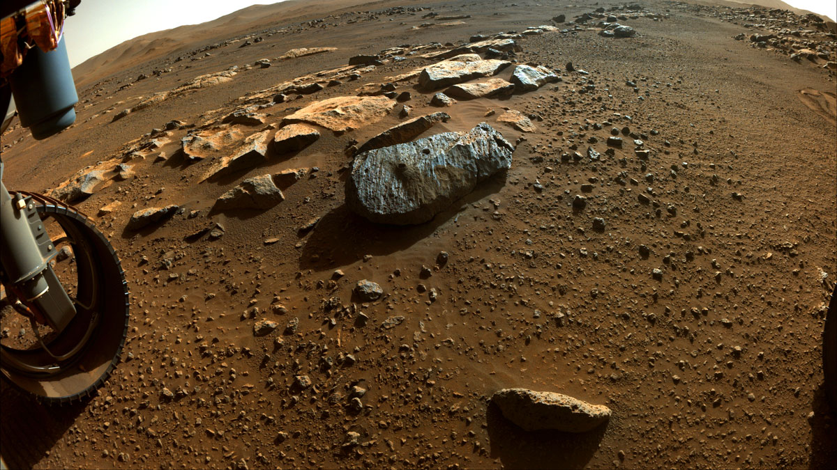 This is a colored image of Perseverance capturing the sandy, rocky surface of Mars. Perseverance’s wheel in the bottom left of the image. There are large boulders and tiny rock particles present in this image.