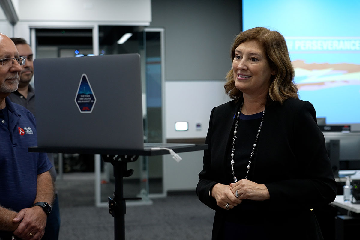 Laurie Leshin, director of NASA's Jet Propulsion Laboratory, smiles at a computer screen to interact with students being honored virtually, with two Mars team members behind the computer monitor, and other team members in the background, near a multi-colored "You've Got Perseverance" graphic on a large screen.