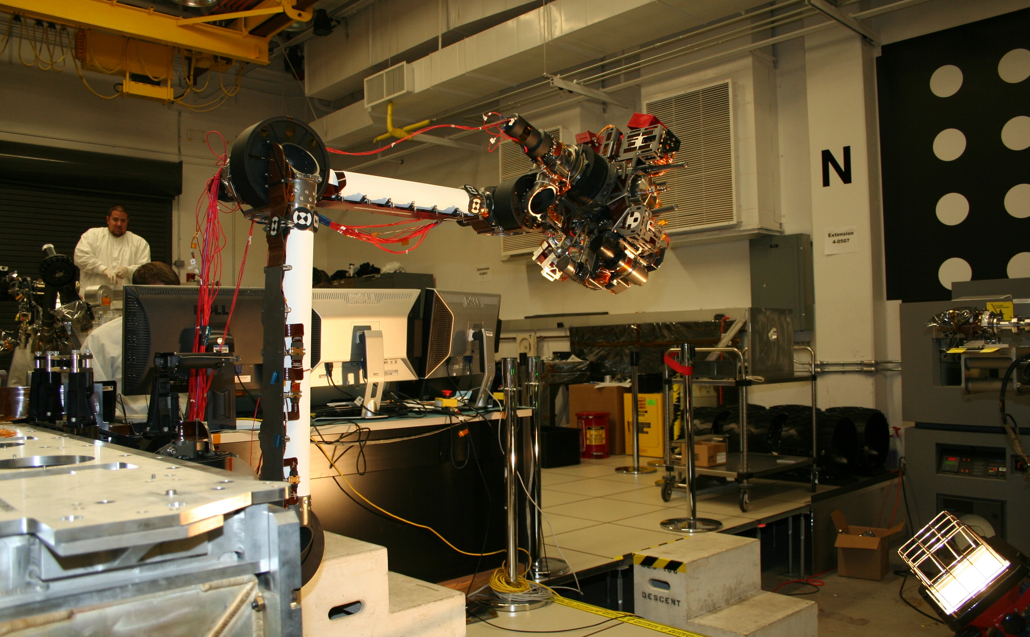 In the middle of this picture, the robotic arm is bent at nearly a 90-degree angle, with the instruments on the end of the arm reaching to the right.  Behind the arm is the laboratory where it is being tested. One engineer is hidden behind a bank of computers, while another with a goatee stands watching in the back, beneath a yellow ceiling crane.  Both engineers wear white lab coats.  Metal lab equipment is scattered throughout the room.