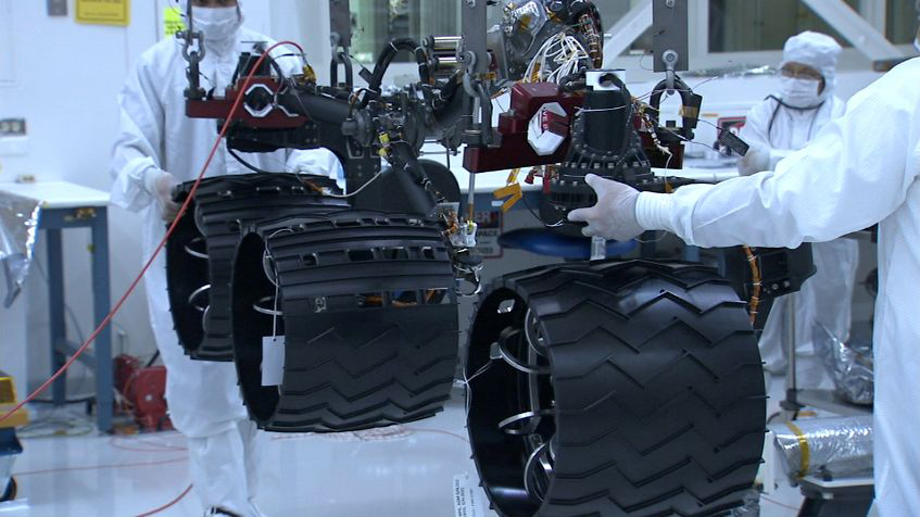 In the middle of this image, three wheels are shown raised by a lift, with engineers on both sides of the wheels in the cleanroom, where the Curiosity rover is being assembled. The wheels are made out of aluminum but have a black coating, which prevents the wheels from slipping.  The wheels are attached to dark poles, which bend like 'joints.'  Several black and red cable wires are visible coming out of the poles.  An engineer stands in the background holding a clipboard and looking closely at the rover.  A desk and various tools are visible behind the wheels on the right.