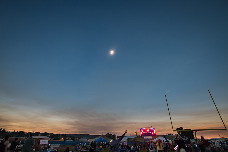 A crowd of people stand at a football stadium. The sky is darkened above them to a dark hazy blue. Near the horizon, the sky is orange. There is a total solar eclipse in the sky.