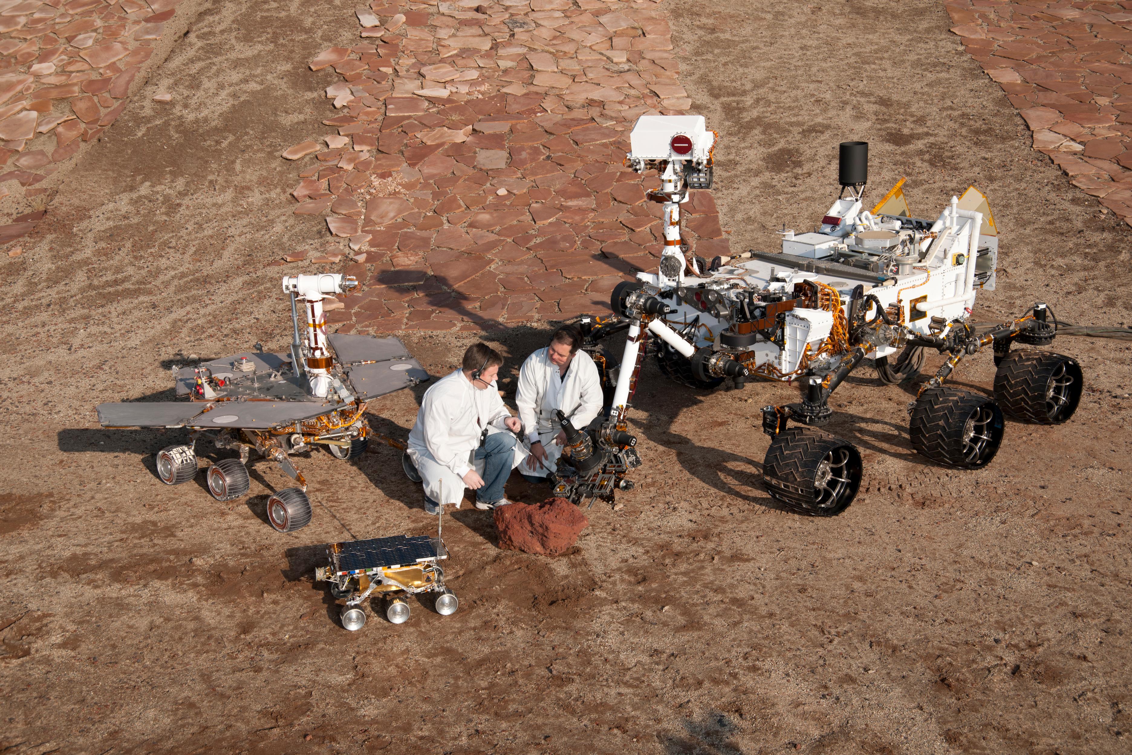 Two spacecraft engineers join a grouping of vehicles providing a comparison of three generations of Mars rovers developed at NASA's Jet Propulsion Laboratory, Pasadena, Calif. The setting is JPL's Mars Yard testing area.
