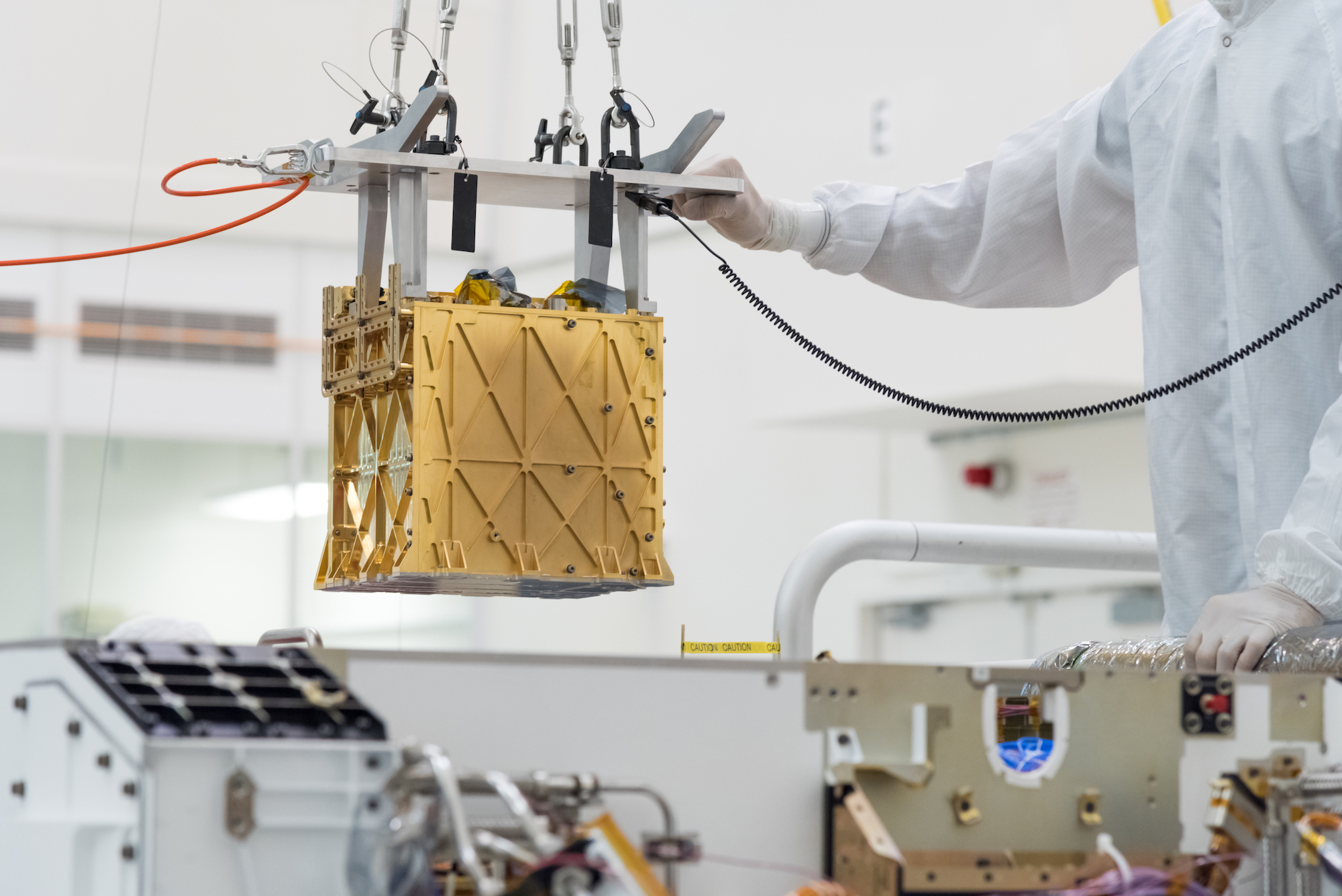 Technicians in the clean room are carefully lowering the Mars Oxygen In-Situ Resource Utilization Experiment (MOXIE) instrument into the belly of the Perseverance rover.