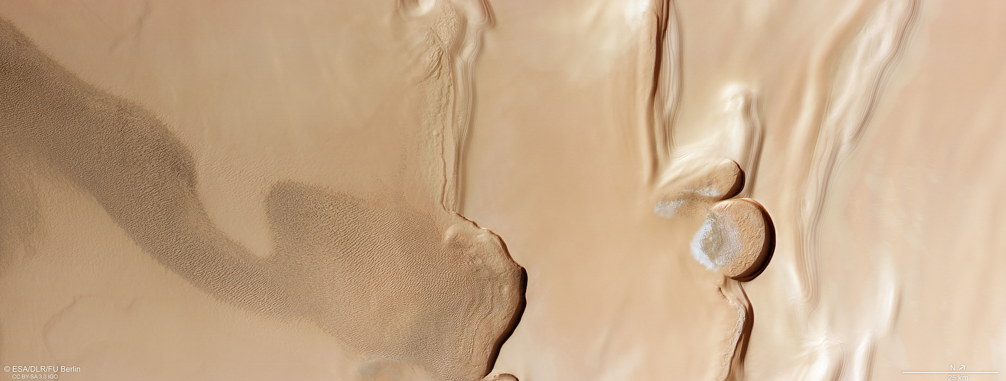 A rectangular slice of tan-colored terrain shows a number of features on Mars: a rippling swath of sand dunes to the left; two notable banks cutting down through the middle of the frame; two steep, semi-circular cliffs in the right-most third of the frame; and smooth terrain to the right. There are signs of layered ground throughout the frame, and the steep cliffs cast dark shadows onto the light orange ground below.