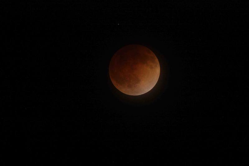 This is an image of the moon during the total lunar eclipse on April 15, 2014. In this image, the moon appears bright orange and is centered on a black sky. Mars is seen a tiny white-red dot on the upper left-hand side of the moon.