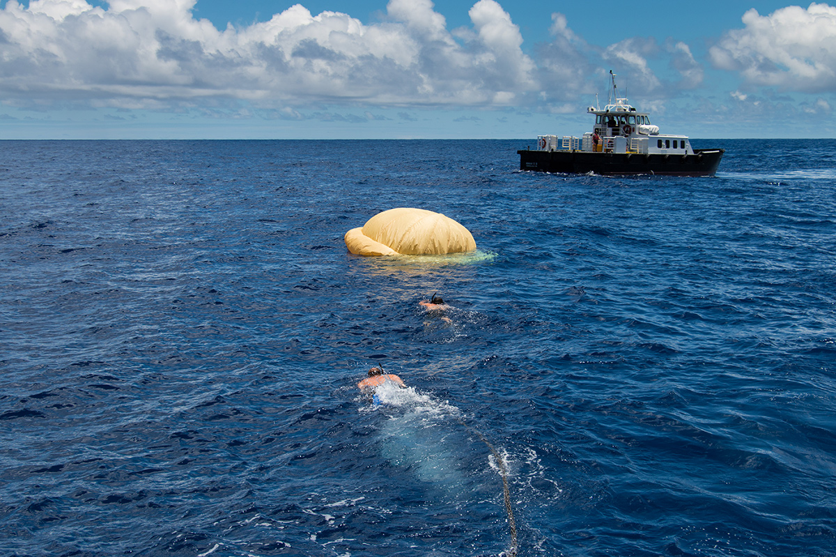Hours after the June 28, 2014, test of NASA's Low-Density Supersonic Decelerator over the U.S. Navy's Pacific Missile Range, two members of the Navy's Explosive Ordinance Disposal swim towards the pilot ballute that was used to deploy the parachute. In the background, the recovery vessel Mana'o II.