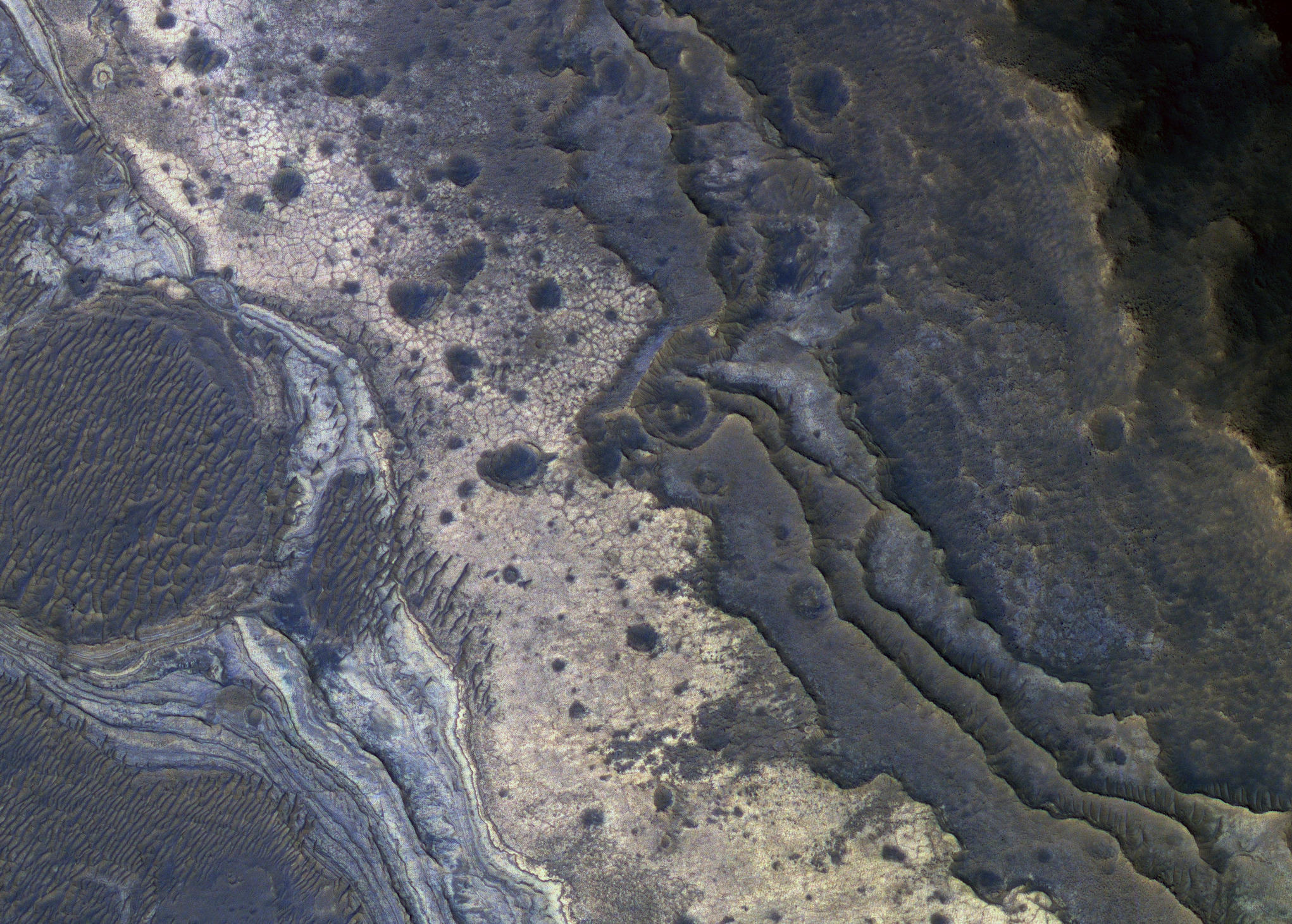 This image shows layers of rock stacked like a staircase, their edges extending diagonally from top left to bottom right and stepping downward from the lower left to the top right. At the far left of the image is an eroded, round crater filled with a row of nearly parallel sand dunes. To the right of the crater, a pinkish-colored layer of light-toned rock within the staircase contains opal.
