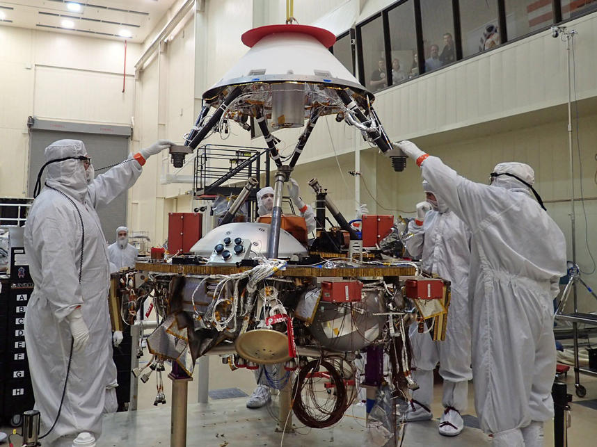 In this photo, spacecraft specialists at Lockheed Martin Space Systems, Denver, are reaching up to guide lowering of the parachute cone for installation onto NASA's InSight spacecraft.