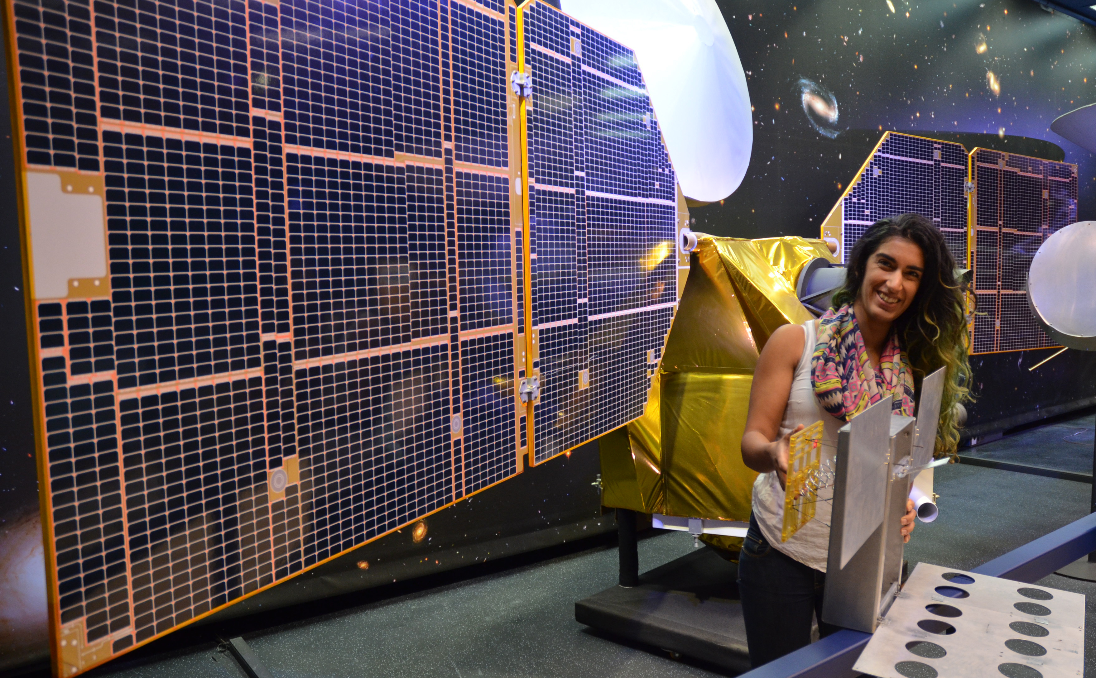 The full-scale mock-up of NASA's MarCO CubeSat held by Farah Alibay, a systems engineer at NASA's Jet Propulsion Laboratory, is dwarfed by the one-half-scale model of NASA's Mars Reconnaissance Orbiter behind her.