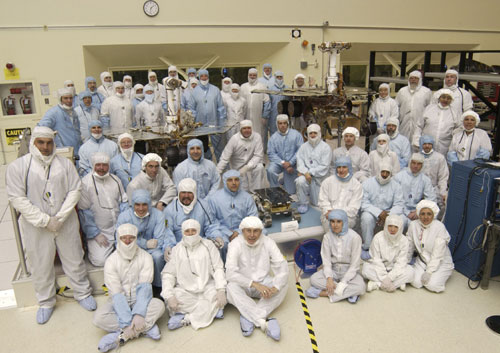 A team of about 50 men and women in white and blue cleanroom smocks and bonnets  stand and sit around their creations: Spirit and Opportunity.  The twin rovers of the Mars Exploration Rover mission are about the size of golf carts.  The significantly smaller, microwave-sized Sojourner rover from the 1997 Pathfinder mission sits in the middle.