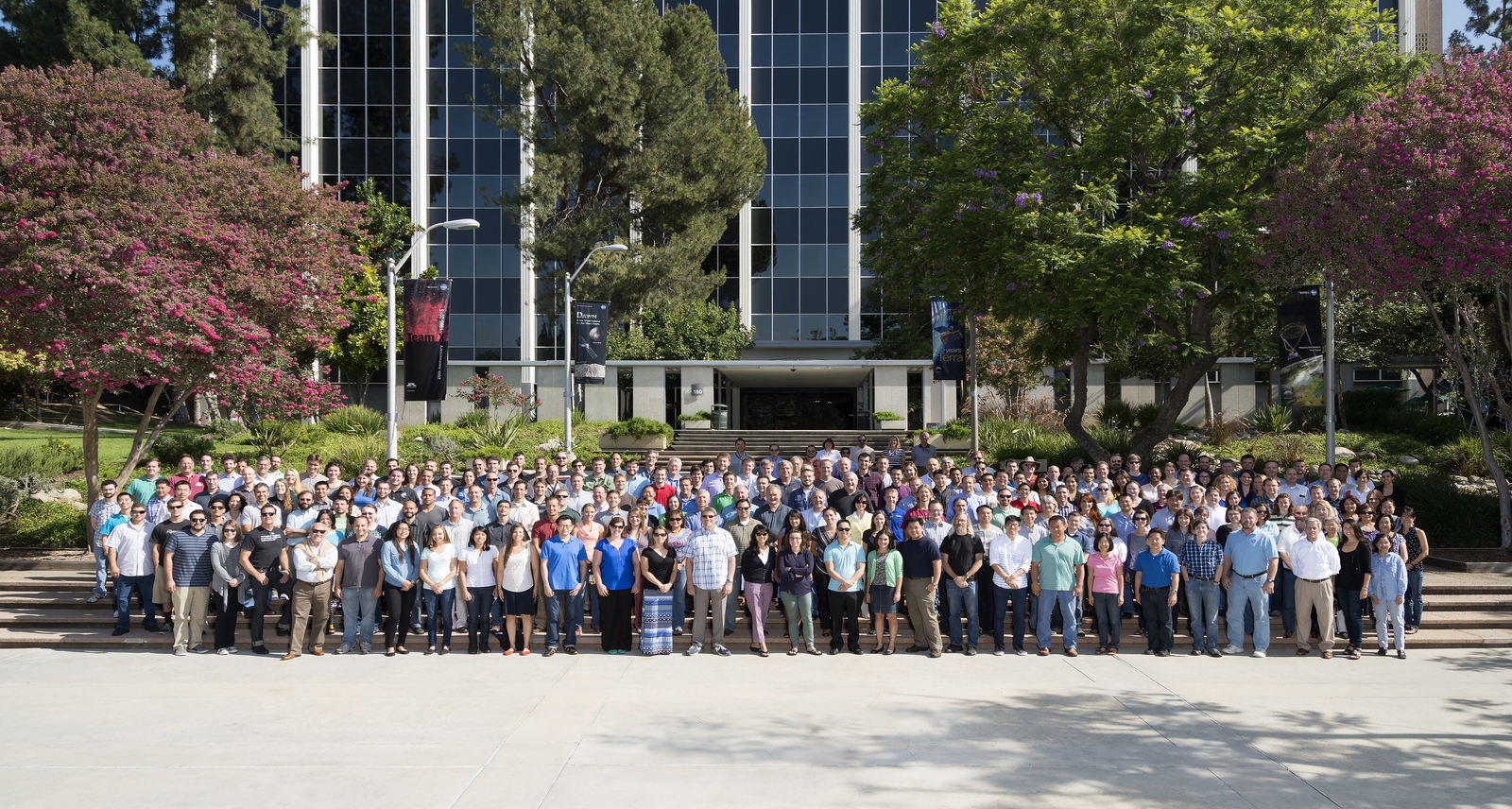 About 100 team members stand in a group photo in front of the steps of a large administration building.